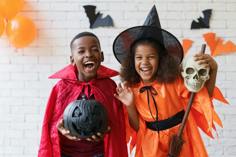Two black children in halloween costumes laughing together in front of decorated brick wall