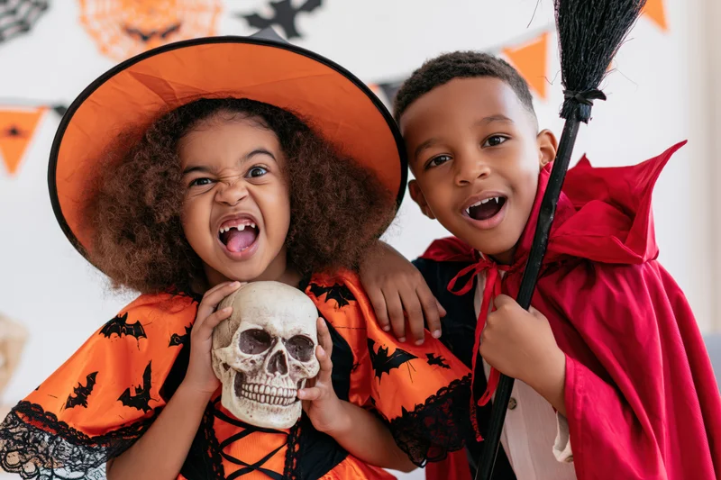 Two black children in halloween costumes posing with skull and broom, smiling and laughing indoors