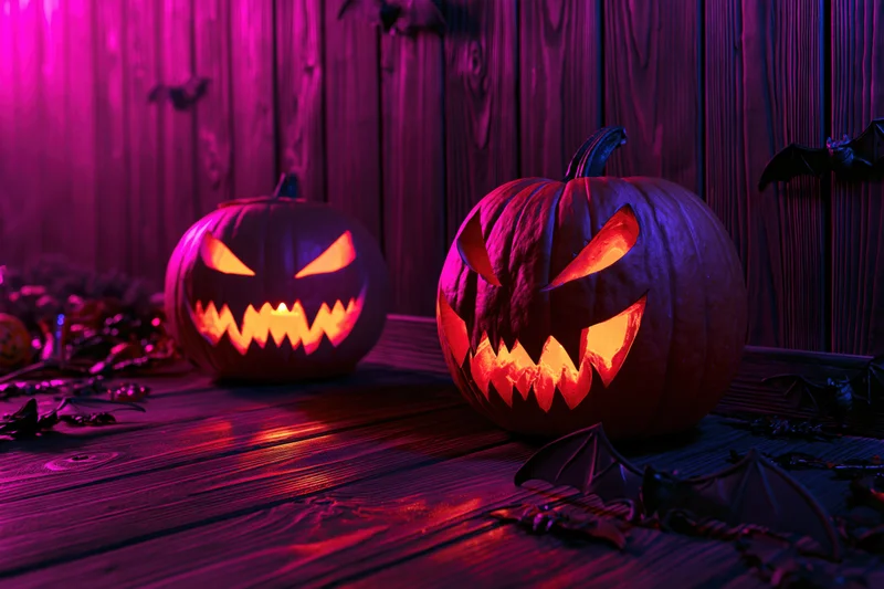 Two carved jack-o-lanterns glowing with orange light on a wooden floor against a dark wooden wall surrounded by bat decorations