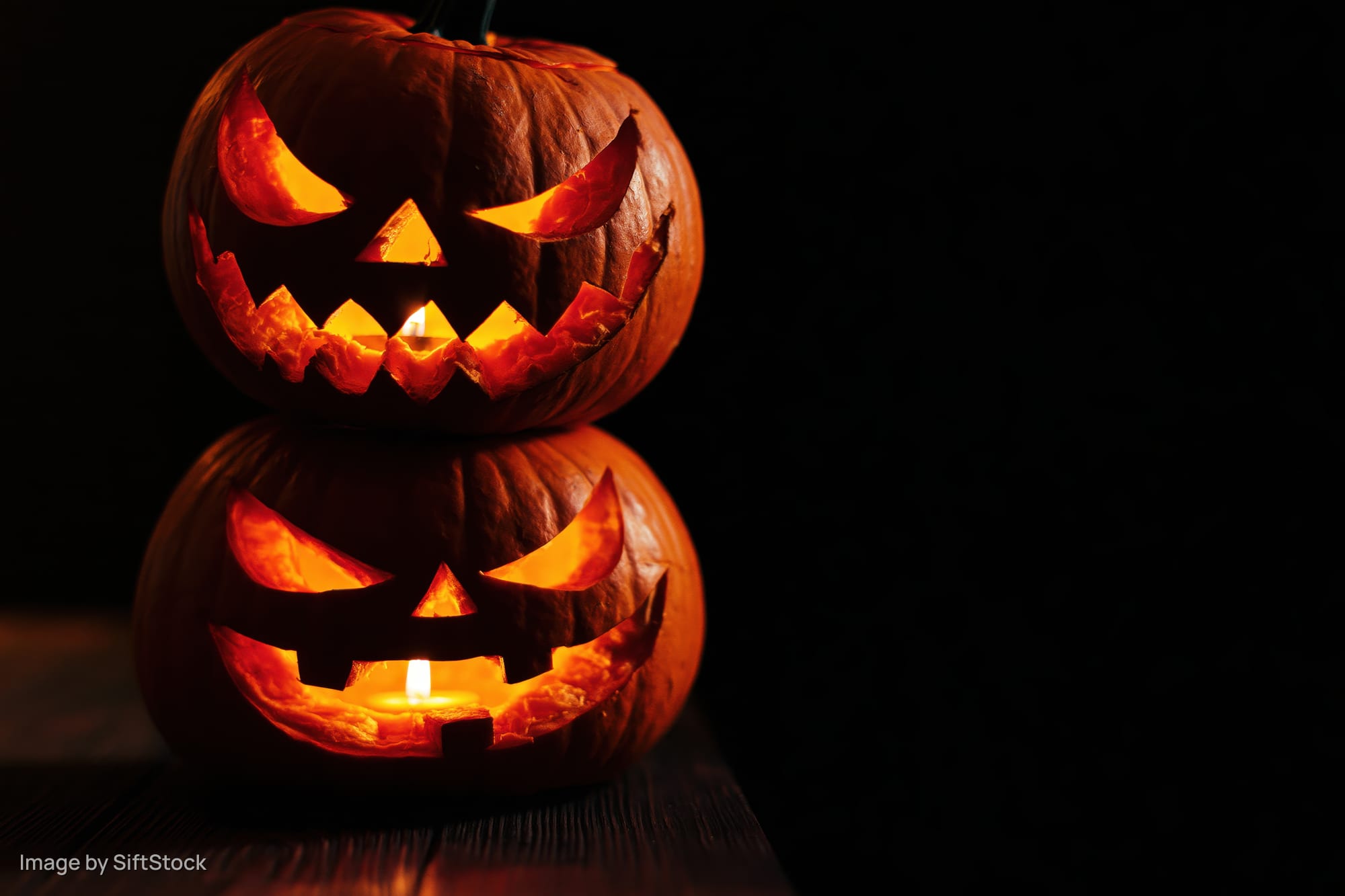 Two carved jack-o-lanterns stacked with candles glowing in the dark on a wooden surface