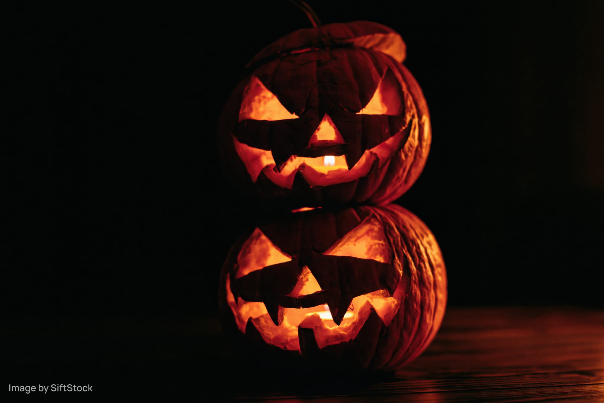 Two carved jack-o-lanterns stacked and glowing with candles on a dark wooden surface