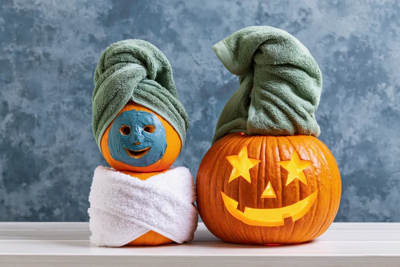 Two carved pumpkins styled as spa characters with towels and facial mask on a table