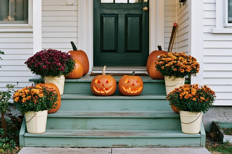 Carved jack-o-lanterns glowing on front porch steps with potted mums and pumpkins