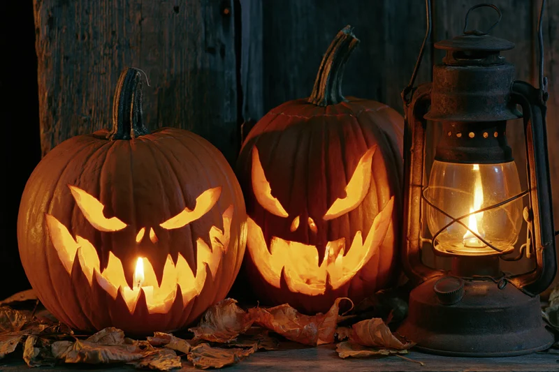 Two carved jack-o-lanterns with glowing faces and a lit vintage lantern on a wooden surface surrounded by dried leaves