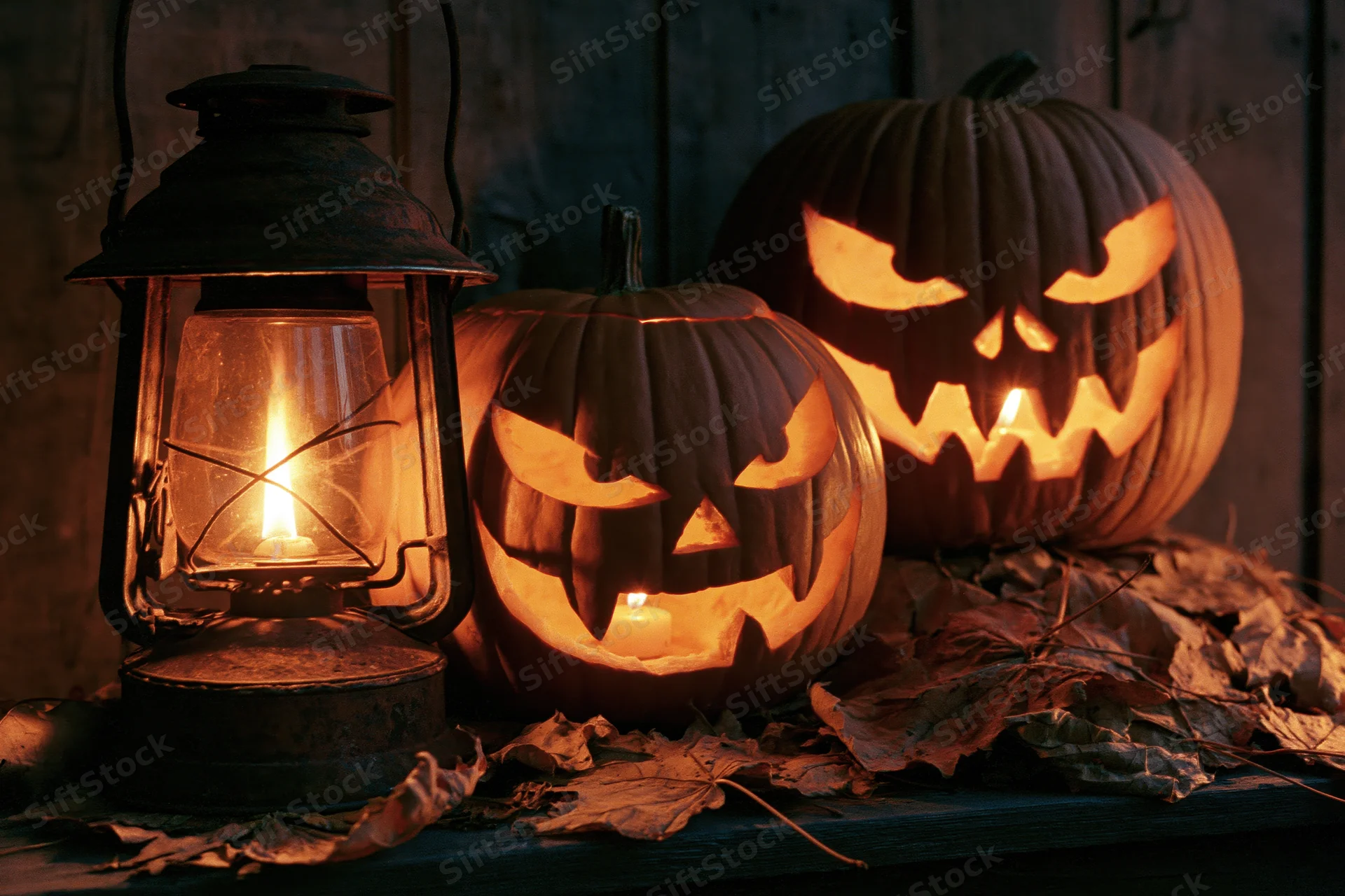 Two carved jack-o-lanterns with glowing faces and a lit lantern on a wooden table surrounded by dry autumn leaves