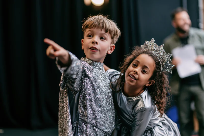 Two children in costumes pointing and smiling on a dark stage during a school play rehearsal