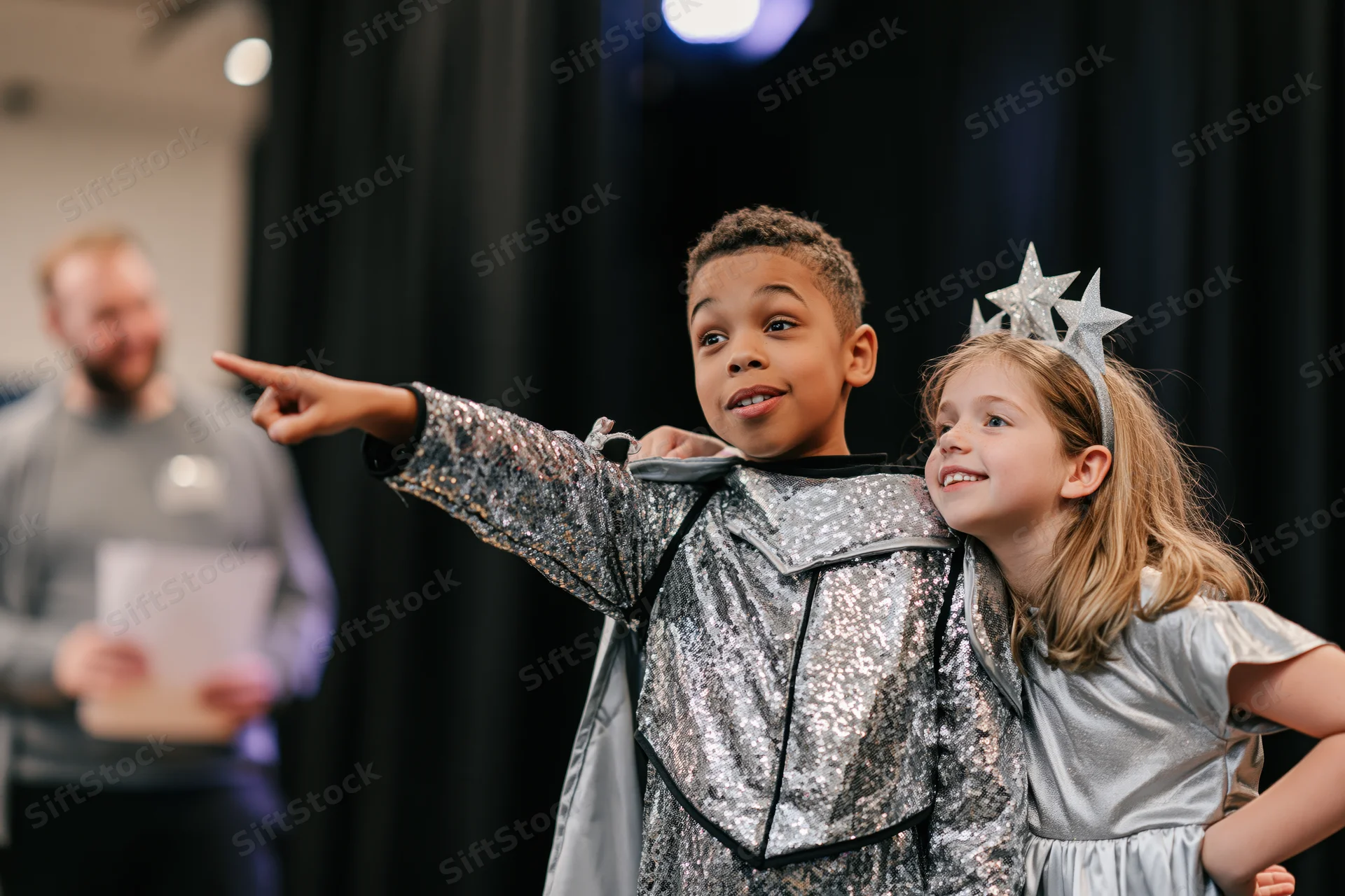 Two children in silver costumes performing on stage, one pointing while the other smiles beside them