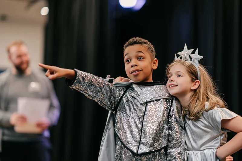 Two children in silver costumes performing on stage, one pointing while the other smiles beside them