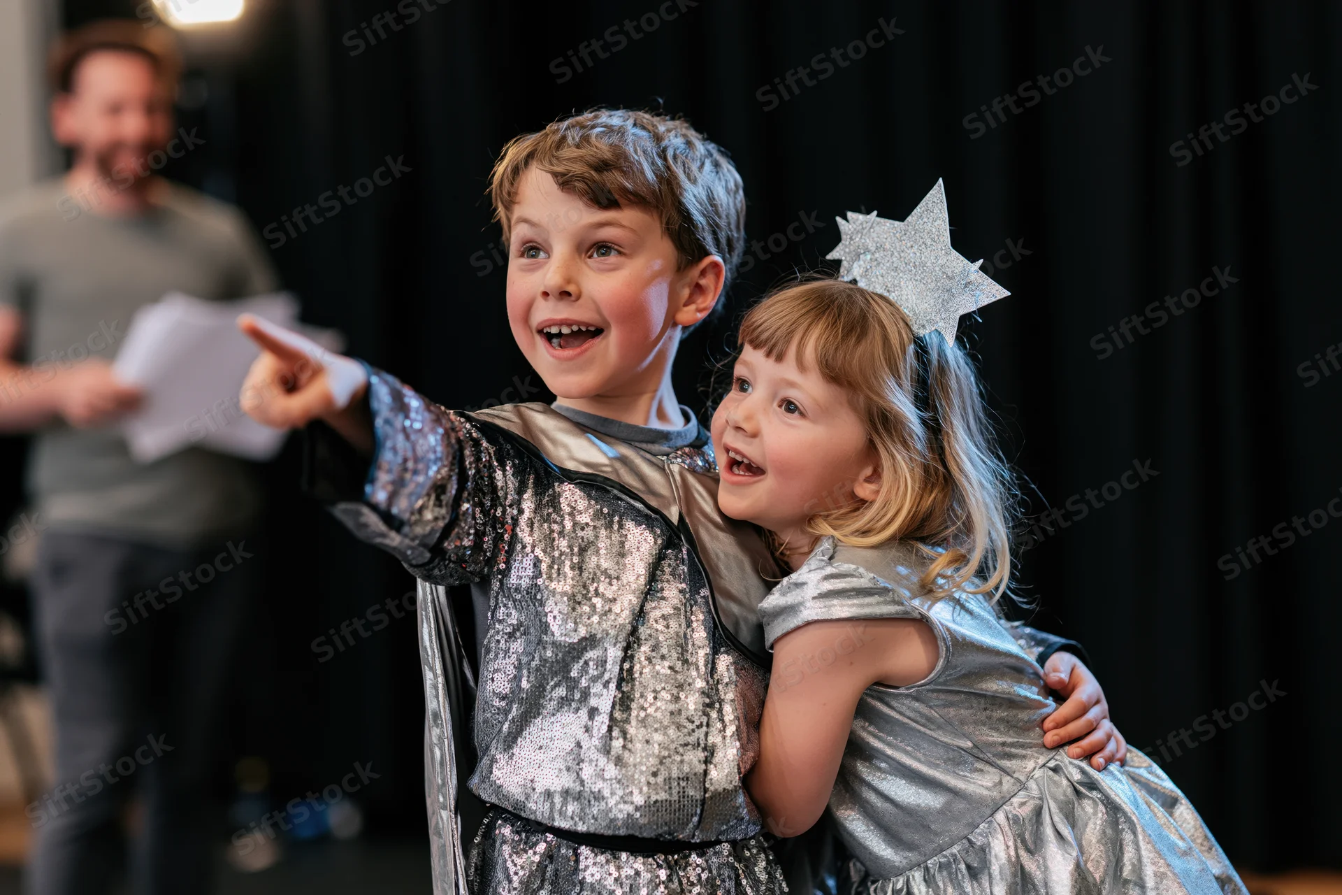 Two children in silver costumes hugging and pointing on a dark stage during a school play rehearsal