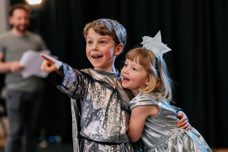 Two children in silver costumes hugging and pointing on a dark stage during a school play rehearsal