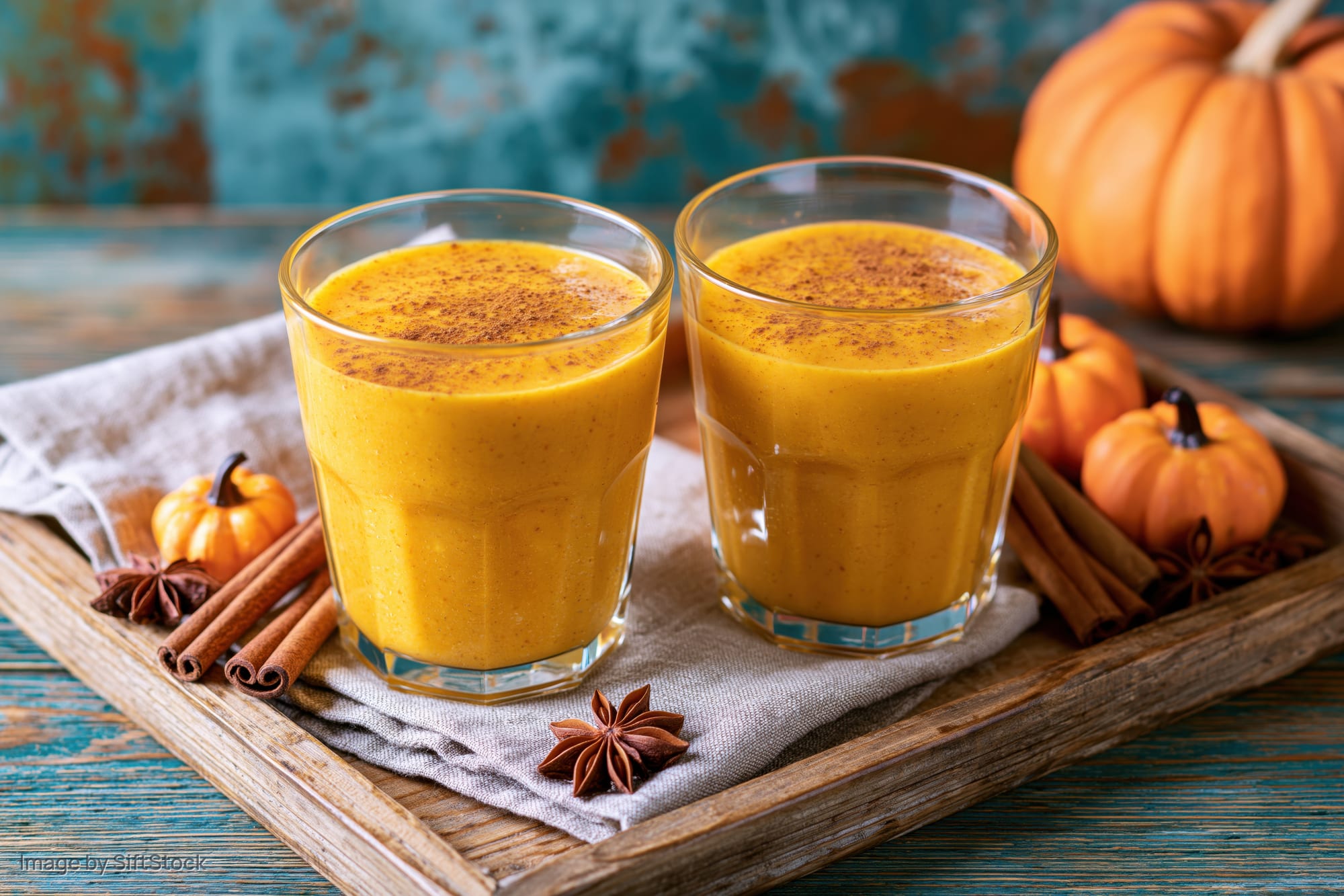 Two glasses of thick orange pumpkin smoothie on a wooden tray with cinnamon sticks and star anise, surrounded by small pumpkins on a rustic table