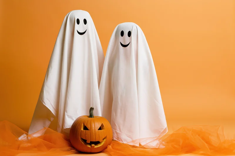 Two smiling ghost costumes made from white sheets standing beside a carved pumpkin on orange tulle against orange backdrop