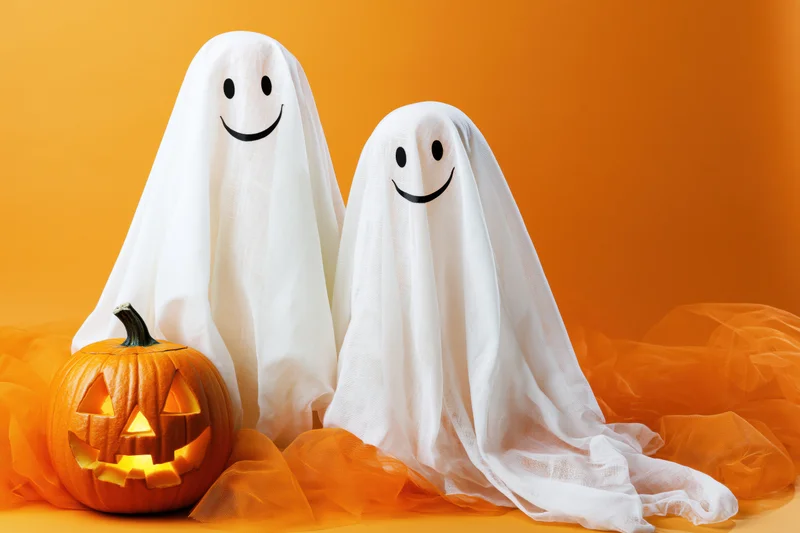 Two smiling ghost costumes made from white fabric sheets beside a glowing jack-o-lantern on orange background