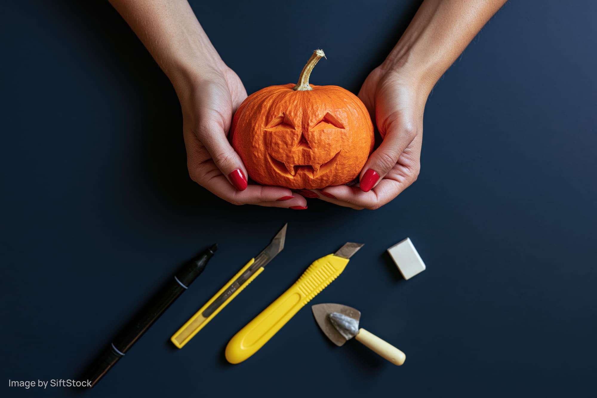 Woman's hands holding a small carved orange pumpkin with carving tools arranged below on dark surface