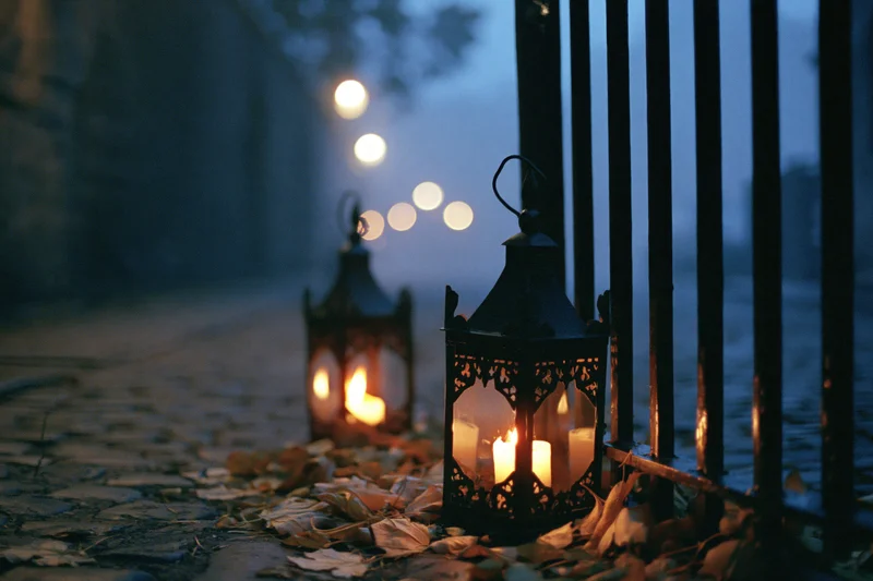 Ornate metal lanterns glowing with warm candlelight on a cobblestone pathway lined with autumn leaves at twilight