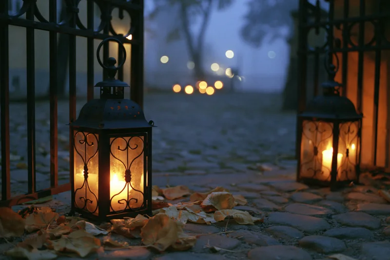 Two ornate metal lanterns glowing with warm candlelight on a cobblestone pathway at dusk surrounded by fallen leaves and a wrought iron gate
