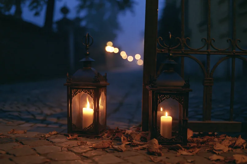 Two ornate metal lanterns with lit candles on a cobblestone path at night near a wrought iron gate surrounded by fallen leaves