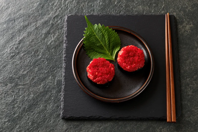 Two red tobiko maki sushi rolls on a black plate with a green leaf and chopsticks on a slate surface