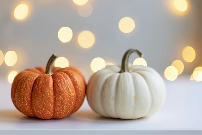 Two small pumpkins, one orange and one white, sitting side by side on a white surface with warm bokeh lights in the background