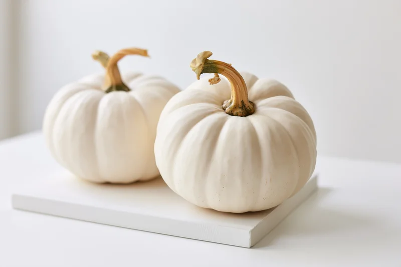 Two small white pumpkins resting on a white board on a clean surface