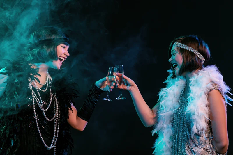 Two women in 1920s flapper costumes toasting with champagne glasses in a smoky, dark setting