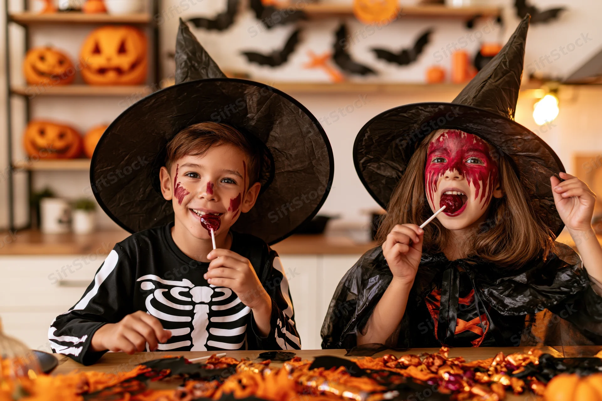 Two children in witch and skeleton costumes eating lollipops at a decorated table indoors