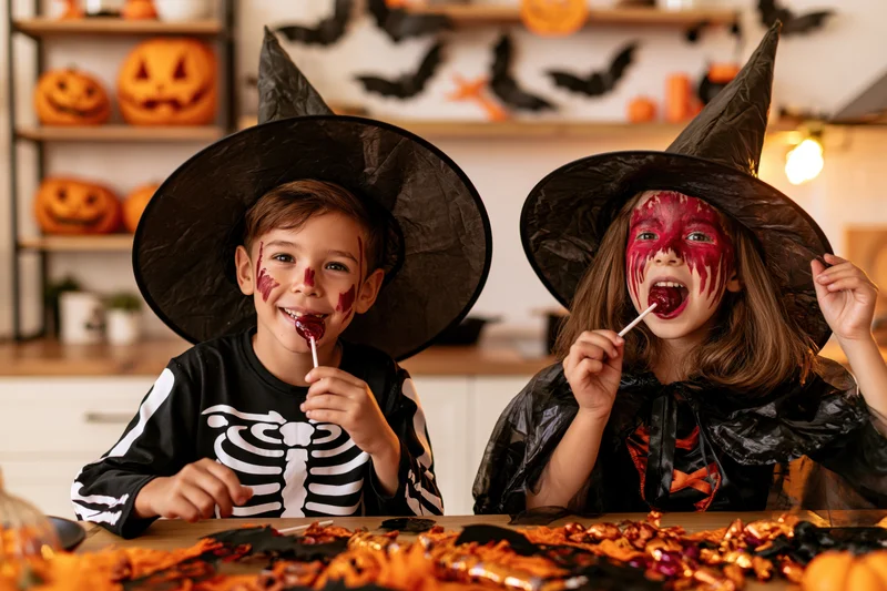 Two children in witch and skeleton costumes eating lollipops at a decorated table indoors