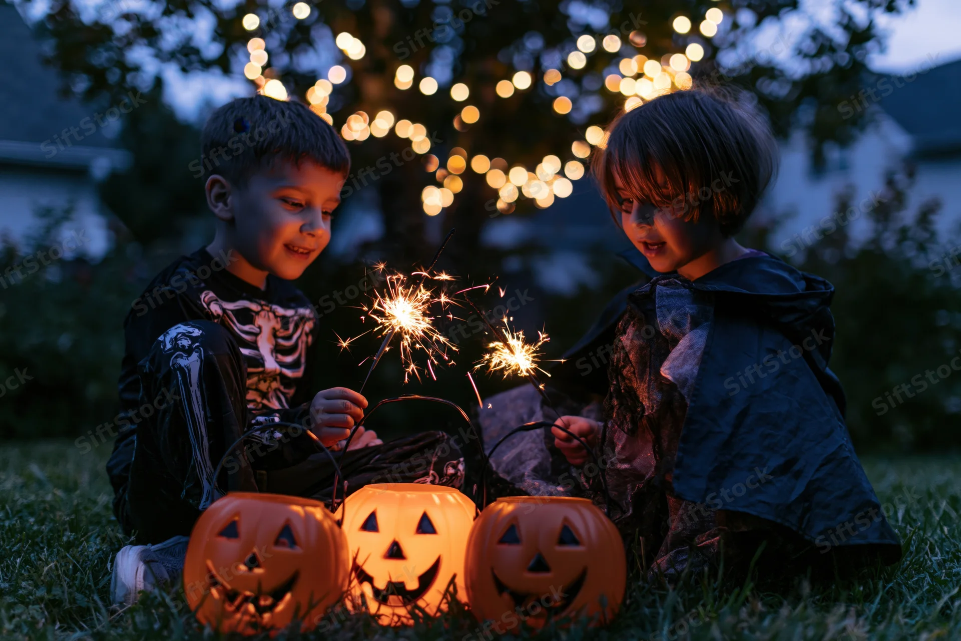 Two children in halloween costumes sitting on grass at night holding sparklers with glowing pumpkins and string lights in background