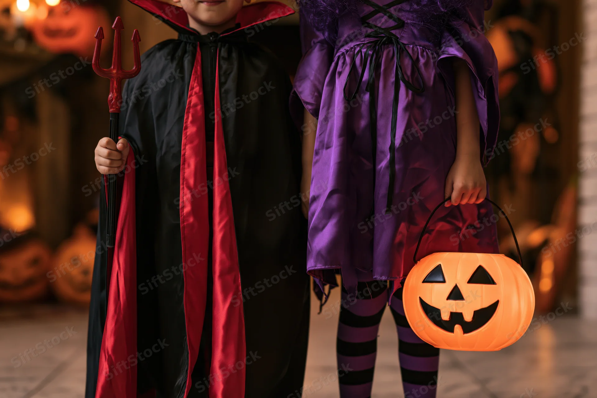 Two children in vampire and witch costumes holding a trident and pumpkin bucket indoors