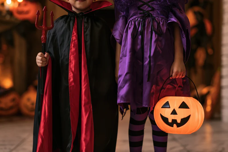 Two children in vampire and witch costumes holding a trident and pumpkin bucket indoors