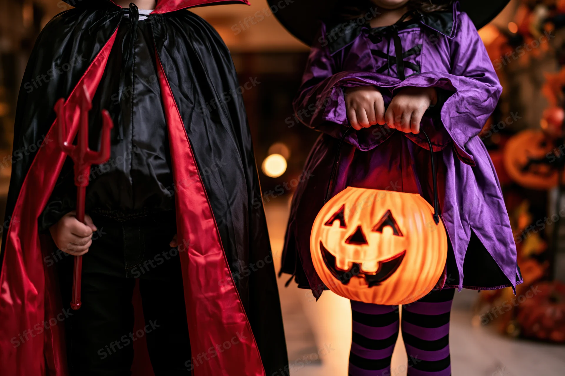 Two children in halloween costumes holding a pumpkin lantern and trident indoors