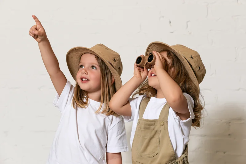 Two young girls playing explorer, one pointing upward, the other using binoculars, both wearing hats against a white brick wall