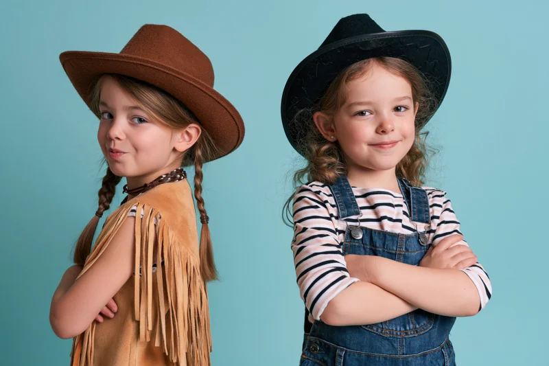 Two young girls in cowboy costumes posing back to back with arms crossed against a turquoise background