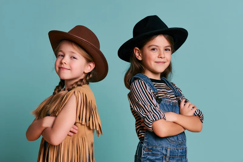 Two young girls in cowboy costumes standing back to back with arms crossed, smiling at camera against turquoise backdrop