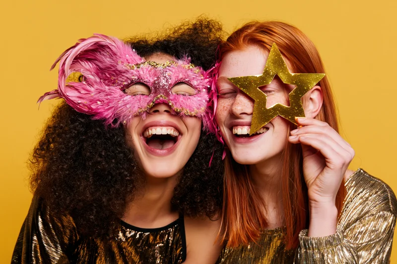 Two young women laughing together wearing carnival masquerade masks against yellow background