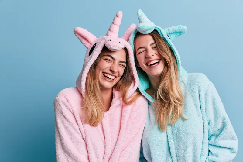 Two young women laughing together in plush unicorn onesies against a blue background