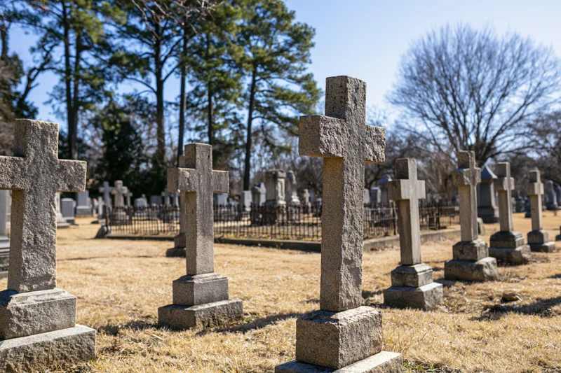 Weathered granite cross grave markers standing in rows across a cemetery with dry grass and trees under a clear blue sky