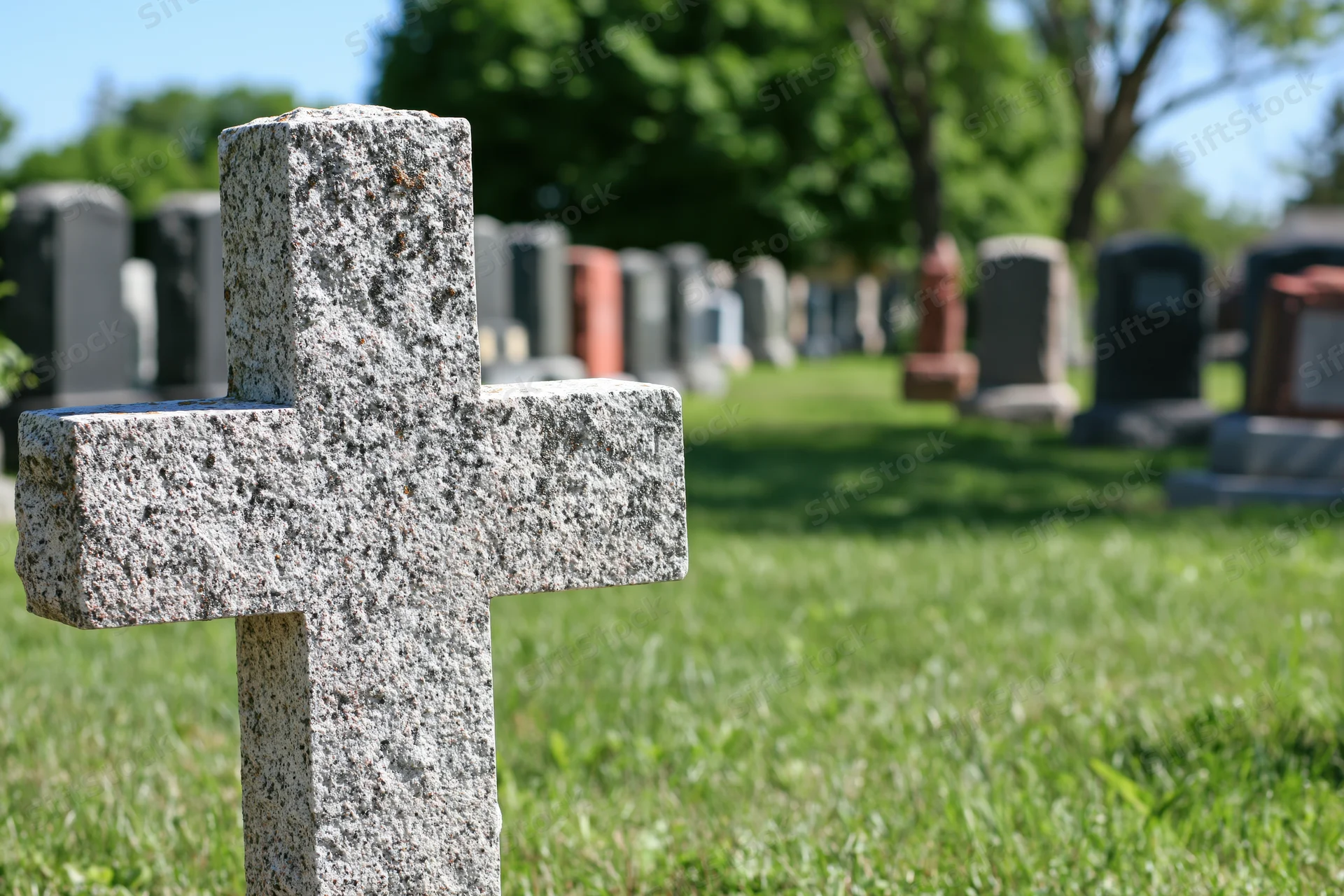 Weathered granite cross gravestone in a grassy cemetery with rows of tombstones in the background