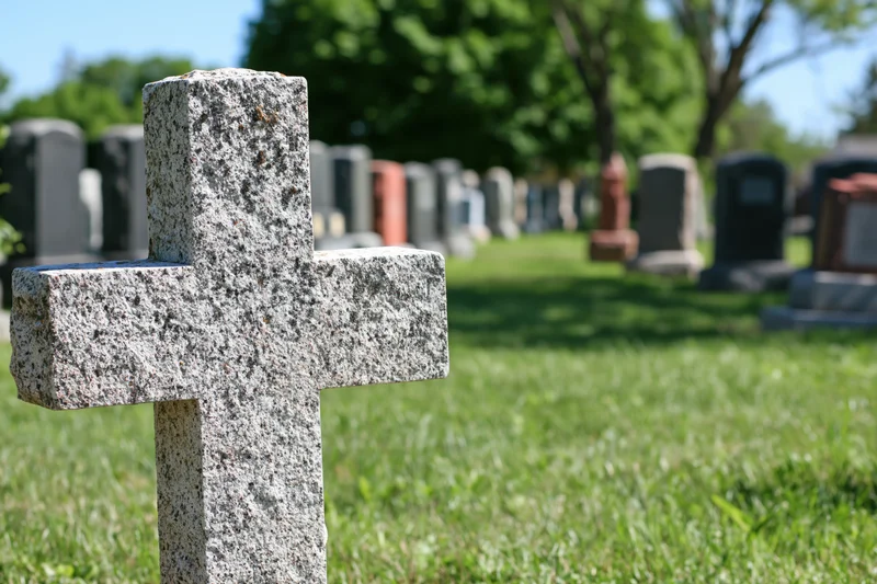 Weathered granite cross gravestone in a grassy cemetery with rows of tombstones in the background