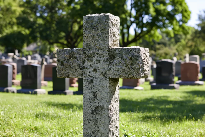 Weathered granite cross gravestone in a grassy cemetery with trees and other tombstones in the background