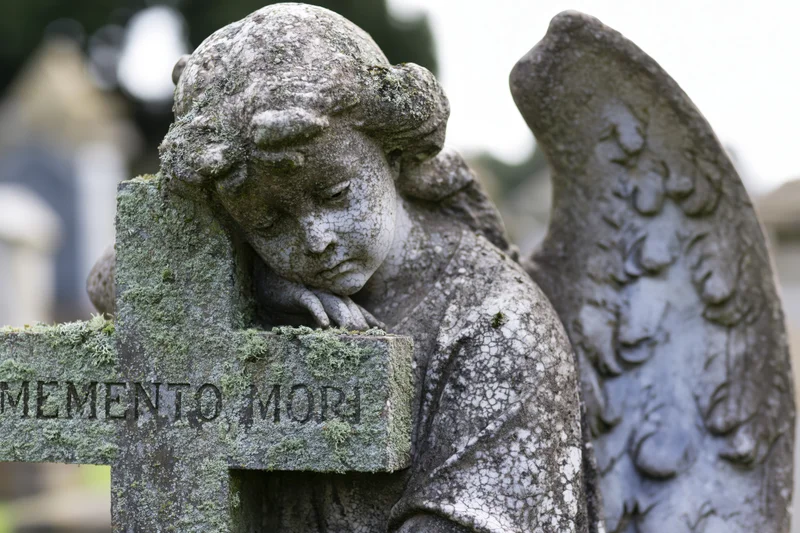Weathered stone angel with downcast eyes leaning on a moss-covered cross in a cemetery