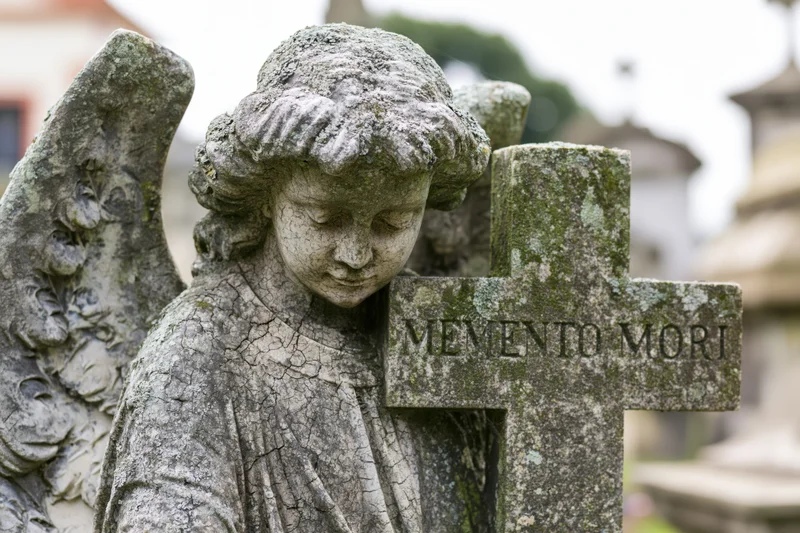 Weathered stone angel sculpture with downcast eyes leaning against a cross inscribed with memento mori in a cemetery