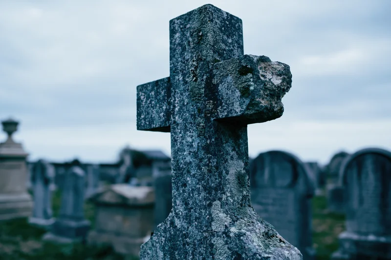 Weathered stone cross gravestone in an old cemetery with blurred tombs in the background