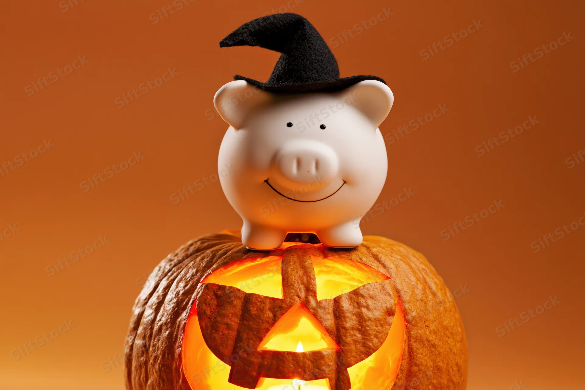 White ceramic piggy bank wearing a black witch hat perched on a glowing carved pumpkin against an orange background