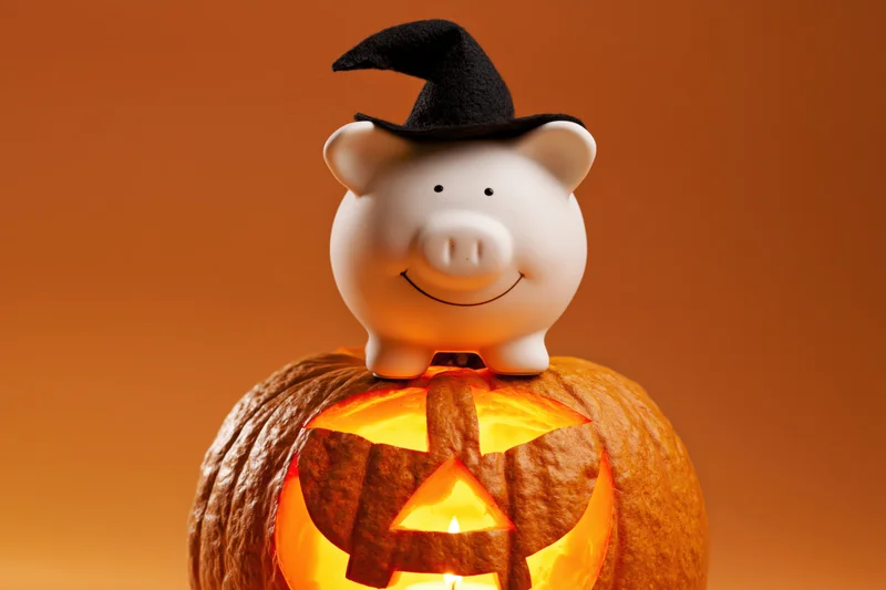 White ceramic piggy bank wearing a black witch hat perched on a glowing carved pumpkin against an orange background