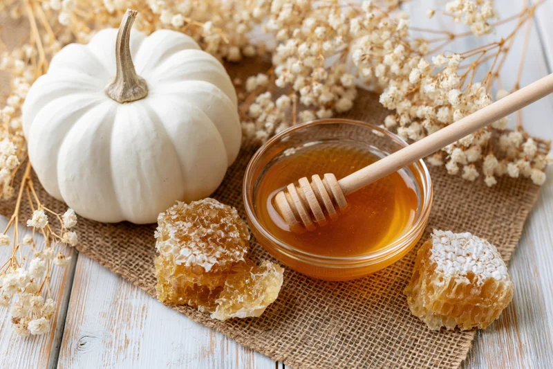 White pumpkin beside glass bowl of honey with honeycomb and dried flowers on burlap over wooden table