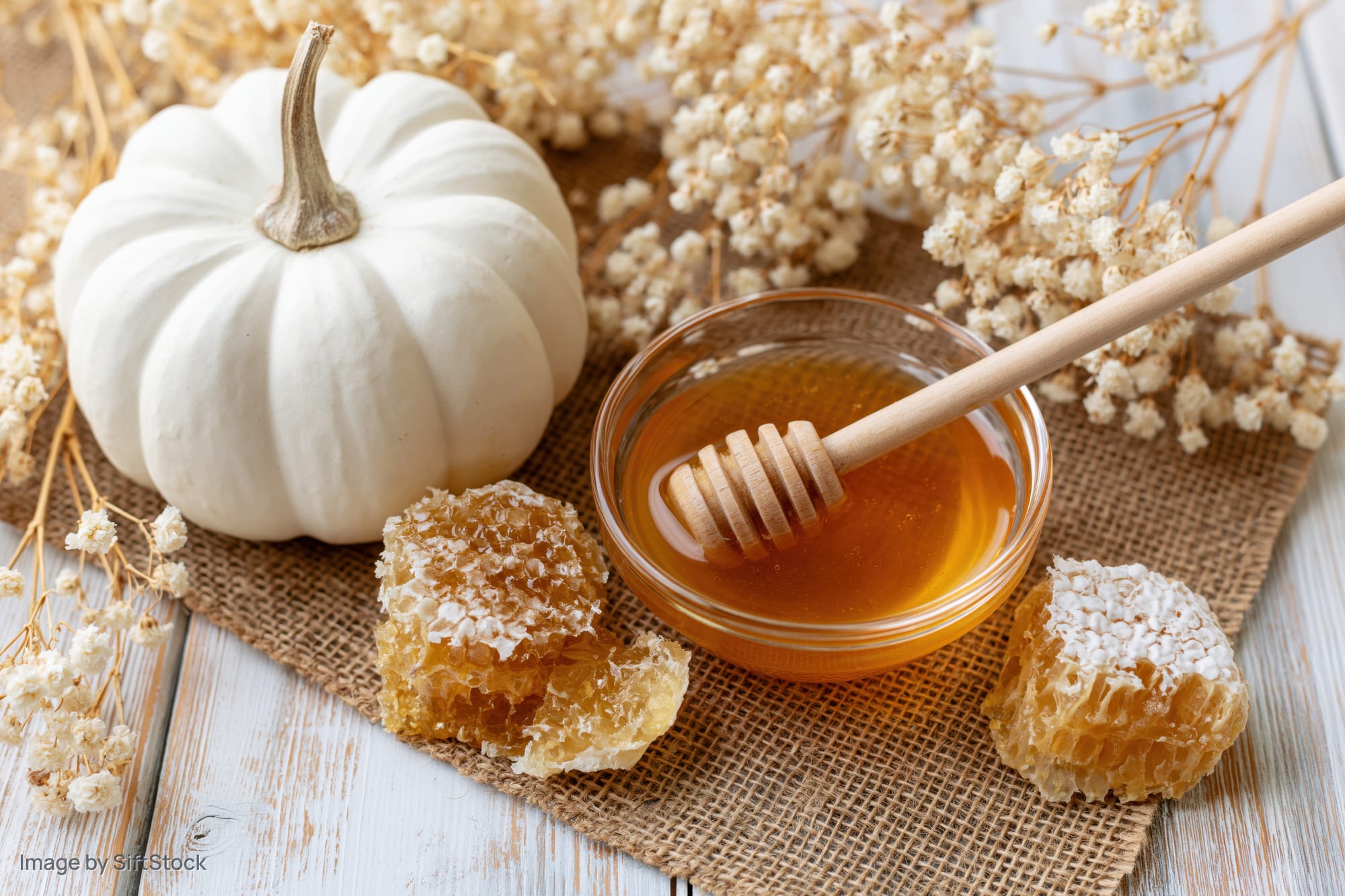 White pumpkin beside glass bowl of honey with honeycomb and dried flowers on burlap over wooden table