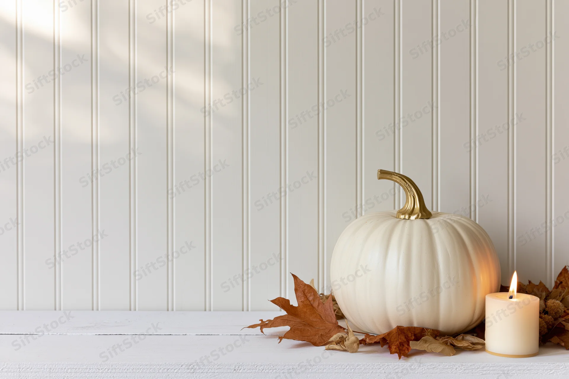 White decorative pumpkin with gold stem and lit candle on wooden table beside autumn leaves against white paneling