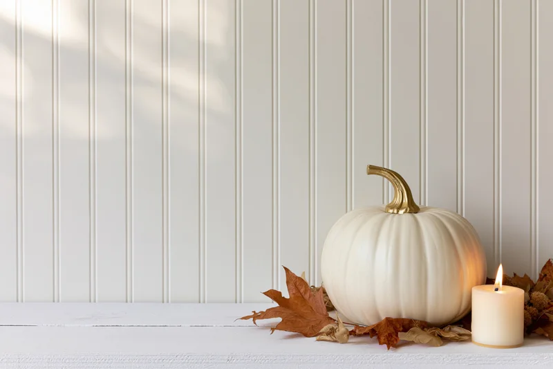 White decorative pumpkin with gold stem and lit candle on wooden table beside autumn leaves against white paneling