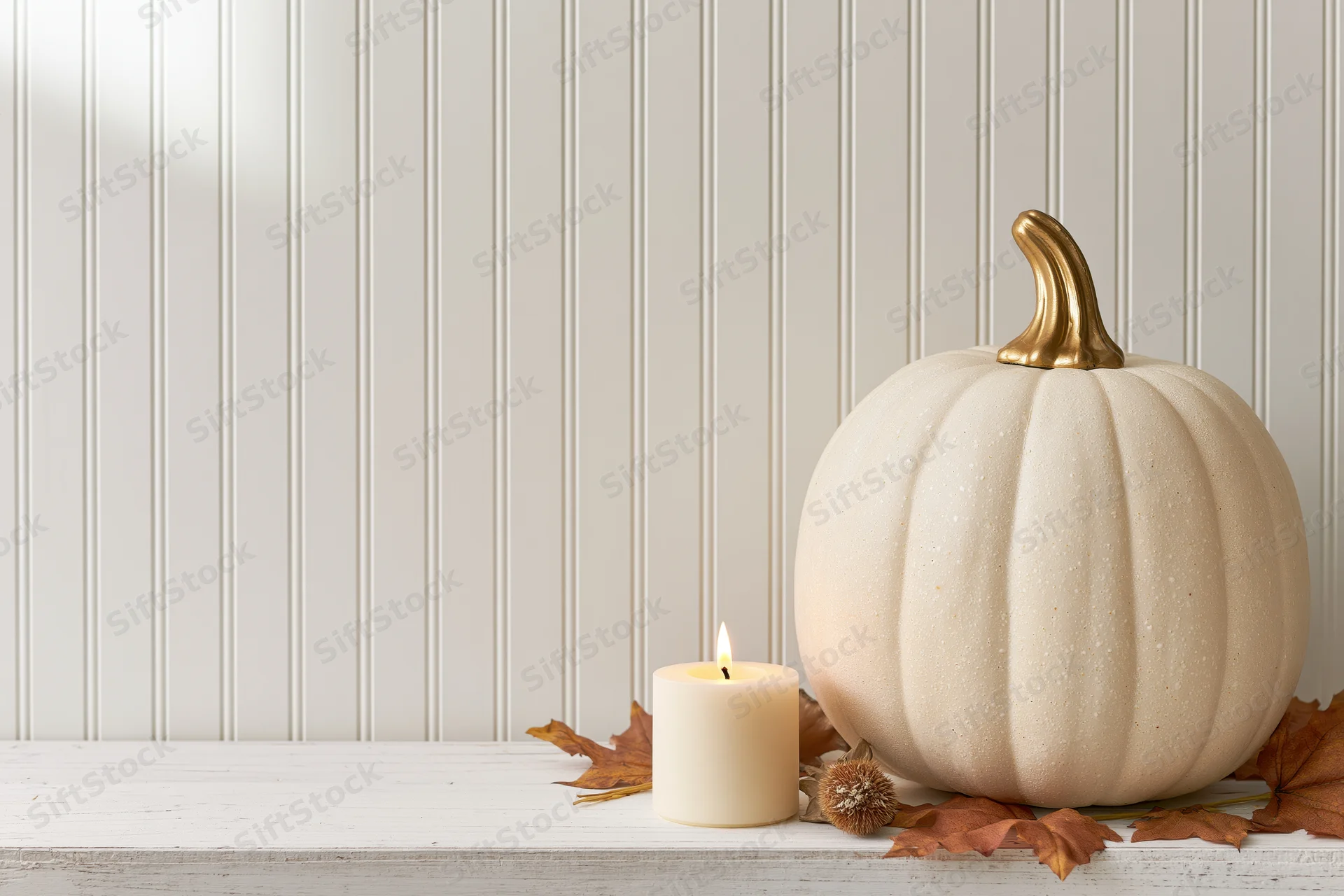 White decorative pumpkin with gold stem and lit candle on a white shelf surrounded by autumn leaves